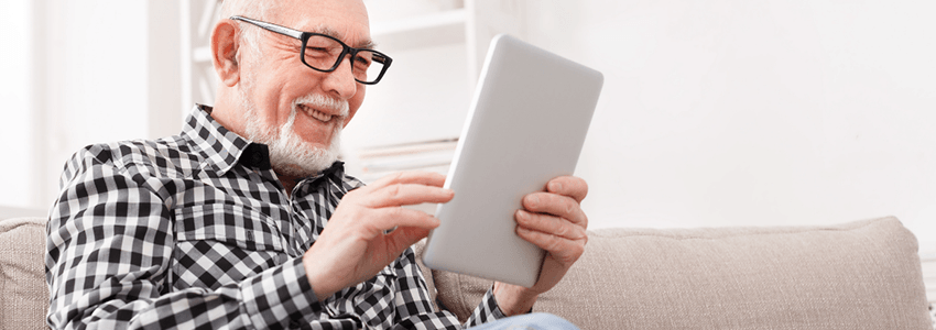 A mature man sits on his couch, smiling, while looking at a tablet.